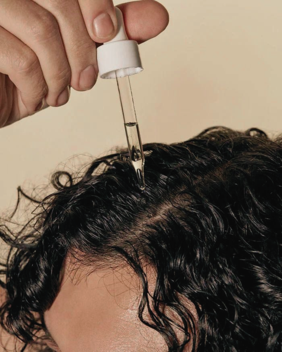 Person applying a dropper of liquid to their hair against a beige background